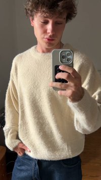 Krém színű, bolyhos, oversized pulóver, ejtett vállakkal és bordás mandzsettákkal, sötét farmernadrággal párosítva. A textúra lágy és melegnek tűnik.