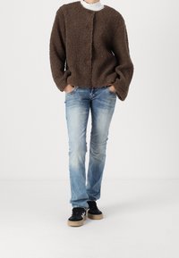 Person som bär en brun fleecejacka, ljusblå jeans, svarta sneakers med vita ränder, med händerna i jackfickorna, stående på ett vitt golv.