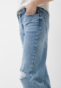 Persona con vaqueros de denim claros y rotos, con la mano en el bolsillo delantero, frente a un fondo claro y liso.