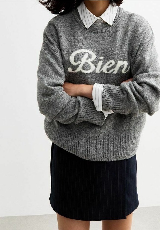 Personne portant un pull gris avec le texte « Bien » sur une chemise blanche à col, associée à une jupe noire à fines rayures, bras croisés.