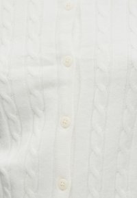 Biely kardigán s pleteným vzorom, ktorý má zapínanie na prednej strane so štyrmi okrúhlymi gombíkmi, mäkkú textúru a výrazné káblové vzory po celej látke.