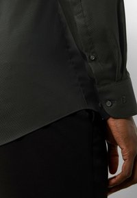 Schwarzes Hemd mit strukturiertem Muster, Knopfmanschetten und abgerundetem Saum; der Stoff wirkt glatt und leicht.