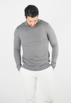 Pull gris à manches longues, col rond, texture lisse, design ajusté, associé à un pantalon blanc. Marquage subtil sur l'ourlet.