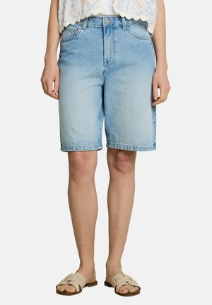 Hellblaue Jeansknielange Shorts, getragen von einer stehenden Person, kombiniert mit beigen Slides und einem gemusterten weißen Oberteil.