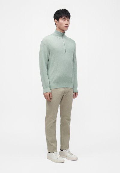 Jeune homme debout portant un pull à fermeture éclair vert clair, pantalon beige et baskets blanches sur fond blanc uni.