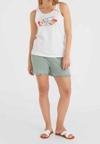 Biały top na ramiączkach z kwiatowym nadrukiem i napisem, zestawiony z jasnzielonymi szortami z falowanym wykończeniem. Modelka nosi białe sandały na płaskiej podeszwie.