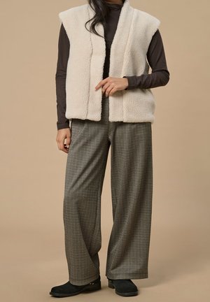 Persoon draagt een mouwloos beige fleecevest over een donkerbruine top met lange mouwen, wijde geruite broek en zwarte schoenen tegen een beige achtergrond.