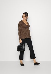 Top de maternidade com um design de drapeado numa padrão floral escuro, combinado com calças pretas largas e sapatos confortáveis pretos. A segurar uma mala estruturada preta.