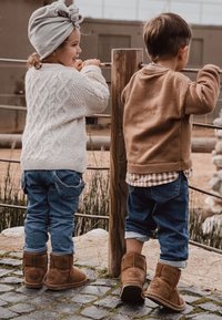 Dwoje dzieci stoi przy drewnianym ogrodzeniu. Jedno ma na sobie biały sweter w warkocze i szary turban; drugie w brązowej polarowej bluzie i koszuli w kratkę, oboje w niebieskich dżinsach i brązowych botkach za kostkę.