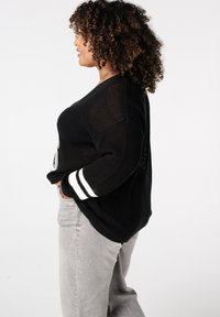 Vrouw met krullend haar, die een zwart opengewerkt truitje met witte strepen op de mouwen en een grijze spijkerbroek draagt, staand in profiel met de handen in de zakken.