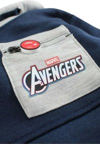 Patch Marvel Avengers sur une poche en tissu gris avec un bouton accent rouge, placé sur un vêtement bleu foncé. Logo de la marque et détails du design clairement visibles.