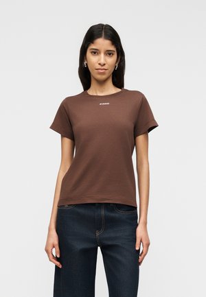 Jeune femme aux cheveux raides et foncés portant un T-shirt marron à manches courtes avec le logo "PINKO" et un jean bleu foncé, debout devant un fond blanc.