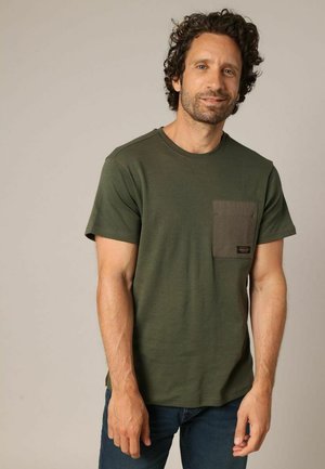 Man met krullend haar, gekleed in een olijfgroene t-shirt met een donkerder borstzakje en blauwe jeans, staand voor een effen achtergrond.