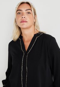 Blusa preta de botão feita de tecido suave, com uma fileira decorativa de lantejoulas prateadas à frente e um colarinho mandarin.