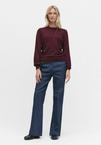Bordowy sweter z długim rękawem i okrągłym dekoltem, zestawiony z ciemnoniebieskimi spodniami w dzwony. Gładka tekstura, dopasowane mankiety i ciemne buty.