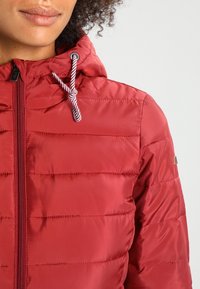 Žena v červené prošívané bundě s kapucí a bílými šňůrkami, zobrazená od ramen až po bradu.