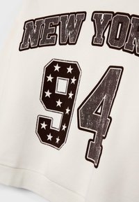 Cream sweatshirt featuring black distressed text "NEW YORK" and the number "94" with star accents. Ribbed hem and soft material texture.