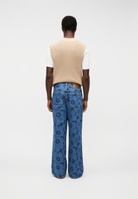 Άνδρας που φοράει μπεζ πλεκτό γιλέκο πάνω από λευκό μπλουζάκι και φαρδιά μπλε τζιν με μεγάλο σκουρόχρωμο λουλουδάτο μοτίβο, στεκόμενος με την πλάτη προς τα έξω μπροστά σε απλό φόντο.