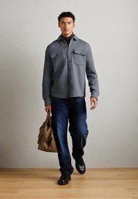 Gray herringbone patterned shirt jacket with two breast pockets, dark jeans, and black shoes, holding a brown leather bag.