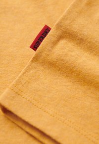 Sennepsgul bomulds T-shirt med en glat tekstur; har et rødt stofmærke broderet med "SUPERDRY" på side sømmen.