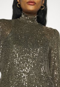 Haut vert olive brillant avec un col haut à volants, présentant un tissu texturé orné de rangées verticales de sequins.