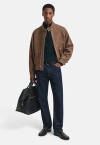 Braune Fischgrätjacke, dunkelgrüner Pullover, dunkelblaue Jeans, schwarze Ledertasche und dunkle Schuhe. Einheitliche Passform mit einem Reißverschlusskragen.