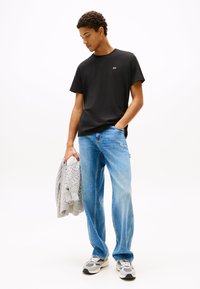 Svart T-shirt med korta ärmar och liten logga, ljusblå jeans med avslappnad passform, och grå stickad tröja, kombinerat med grå och vita sneakers.