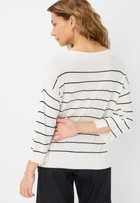 Pull en tricot blanc avec des rayures horizontales noires. Il présente des épaules tombantes et une coupe décontractée. La vue de dos montre un design simple et minimaliste.
