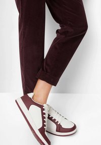 Dolní části nohou v burgundských kordových kalhotách a bílých teniskách s burgundskými akcenty, na jednobarevném světlém pozadí.