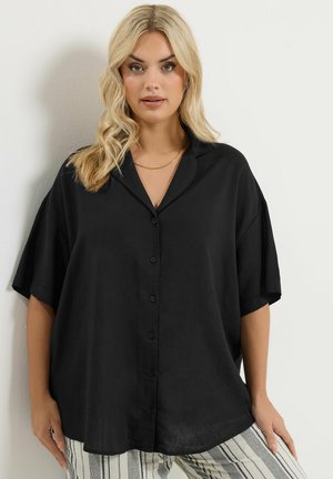 Blonde vrouw die een losse zwarte blouse met knopen draagt en gestreepte lichte broek, staand tegen een effen lichte muur.