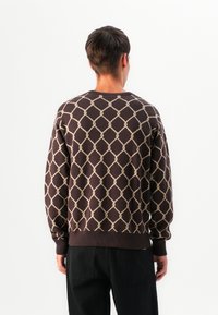 Man wearing a dark brown sweater with a beige diamond pattern, standing facing away against a plain white background.