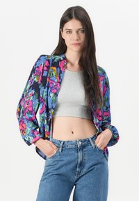 Chemise à boutons florale avec des motifs multicolores en bleu, rose et vert, portée sur un top gris court avec un jean dans une coupe taille haute bleue.