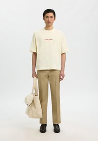 Mladić u svijetlom majici "HOTEL AMOUR", bež hlačama, tamnim cipelama, drži veliku bež torbu s kapom na jednostavnoj bijeloj pozadini.