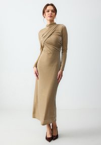 Robe longue ajustée en tissu métallisé doré avec des manches longues et un col haut. Présente des détails drapés sur le devant.
