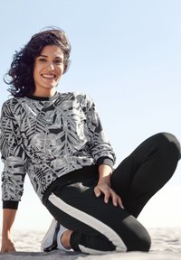 Zwarte en grijze sweatshirt met bladerenpatroon, ronde hals; zwarte joggers met witte zijstrepen, zachte stof, gezeten op een zandige ondergrond.