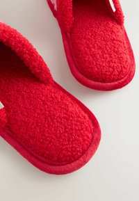 Červené plyšové papuče s textúrovaným látkovým zvrškom, zaobleným dizajnom špičky a mäkkou podšívkou. Okraje olemované hladkou červenou látkou pre zvýšený komfort.