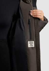 Pufferjack voor in donkergrijs stof, met een rits binnenzak en een label waarop staat "Een warme knuffel van stof", met een soepele textuur.