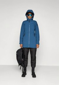 Blue waterproof jacket with a hood, front zipper, and side pockets, worn with black pants and carrying a black backpack.