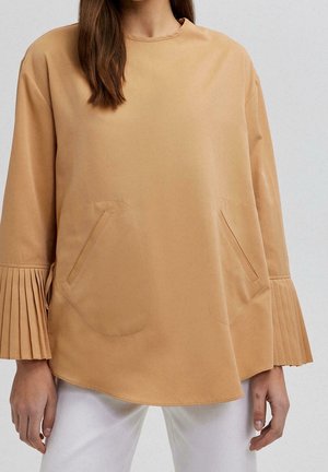 Frau trägt ein lockeres, kamelfarbenes Oberteil mit plissierten Ärmelendes und schrägen Fronttaschen, kombiniert mit weißen Hosen vor einem einfarbigen Hintergrund.