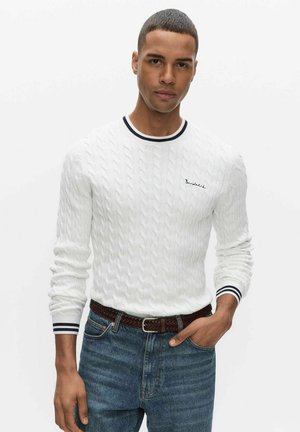 Jeune homme aux cheveux courts portant un pull blanc à torsades avec des bordures bleu marine et un jean bleu, une main dans la poche, sur fond blanc.