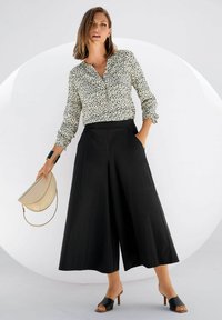 Witte blouse met zwart luipaardprint, gecombineerd met zwarte high-waisted wijde broek. Houdt een beige halfmaanzak vast en draagt zwarte sandalen met hakken.