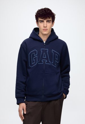Ung mand med mørkt krøllet hår iført en marineblå hættetrøje med lynlås og GAP-logo samt brune bukser, stående mod en helt hvid baggrund.