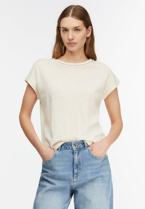 Donna con capelli castani lisci che indossa una maglietta crema a maniche corte e jeans blu a vita alta con le mani nelle tasche su sfondo bianco.