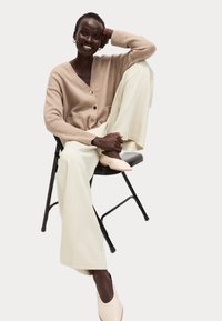 Beige gebreide cardigan met gouden knopen, crèmekleurige wijde broek en blush puntige schoenen. Model zit op een zwarte opvouwbare stoel.