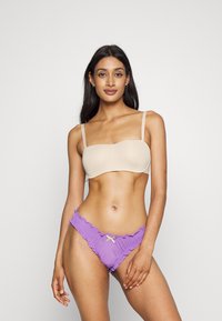Béžová podprsenka bez ramínek s hladkým materiálom je spárovaná s fialovými bikiny s volánmi, ktoré majú dekoratívny žltý akcent v strede.