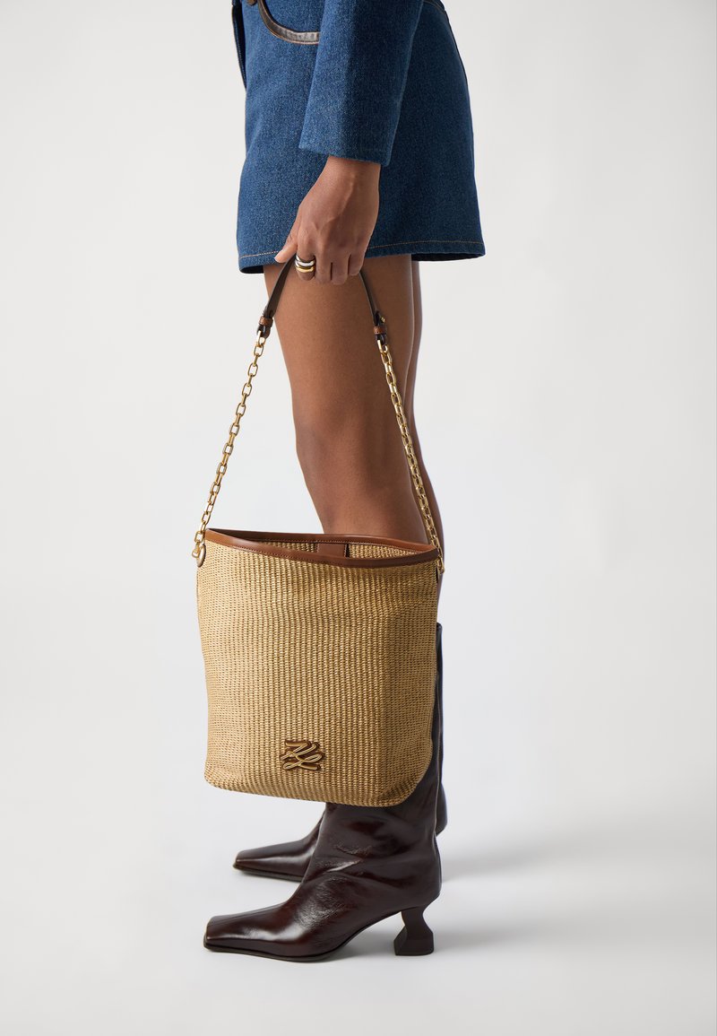 Woven straw bucket bag with brown leather accents, gold chain strap, and a signature logo detail at the front. Bag has an open top.