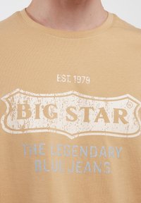 Bežinis medvilnės marškinėlis su pažeistu baltos ir šviesiai mėlynos spalvos grafiniu atspaudu su užrašais "BIG STAR" ir "LEGENDINIAI MĖLYNI DŽINSAI". Apvalus kaklas.