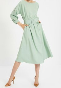 Miętowa sukienka o luźnym fasonie, z rękawami do trzech czwarte, szerokim paskiem w talii i bocznymi kieszeniami, zwiewna spódnica, w połączeniu z cielistymi szpilkami.