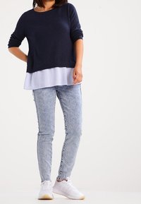 Navy knit sweater over a light blue striped shirt, paired with light-wash skinny jeans and white sneakers with beige soles.