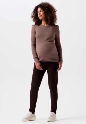 Bruine geribbelde lange mouwen top met pofschouders, gecombineerd met donkerbruine tapered broek en lichte sneakers, gedragen door een zwangere model.